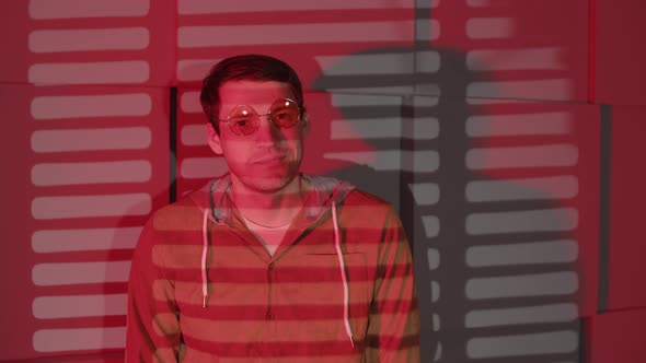 Positive Man in Glasses Standing Near Wall in Dark Room with Falling Red Light and Shadow Blinds alt