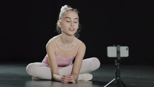 Caucasian Girl Child Teen Ballerina Gymnast Sitting on Floor in Dance Hall Waving Hello to Mobile alt
