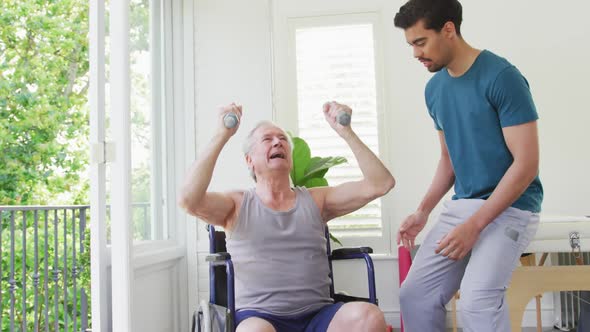 Biracial male coach assisting disabled senior man exercising with dumbbells at fitness studio alt