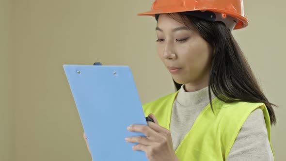Asian Builder Woman Wearing Protective Clothing and a Helmet is Writing Down Something