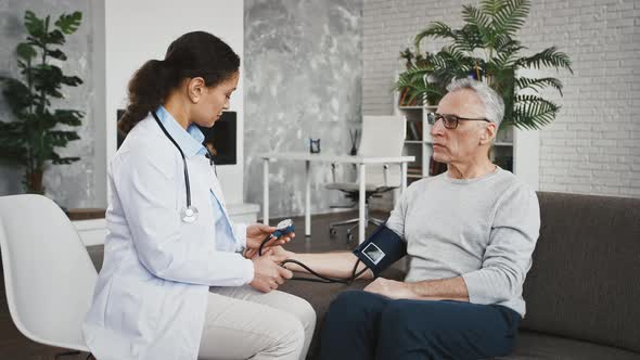 Woman Doctor Measuring Blood Pressure of an Aged Man with a Bloodpressure Cuff Sitting on Sofa alt