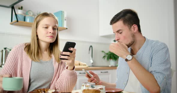 Busy Young Couple Use Smartphones Eating Breakfast in Kitchen at Home Office