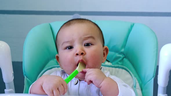 A hungry baby is gnawing on a plastic spoon at the table on a high chair. Teething, whims, itchy gum alt