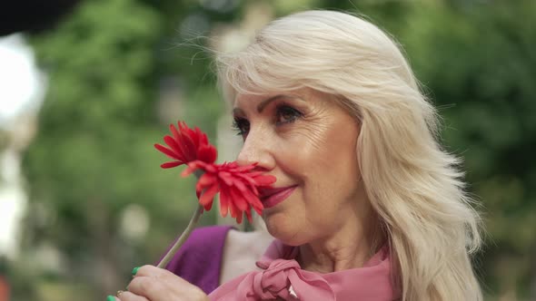 Closeup Blond Adult Woman Smelling Red Flower Smiling Looking Up Standing in Summer Park Outdoors alt