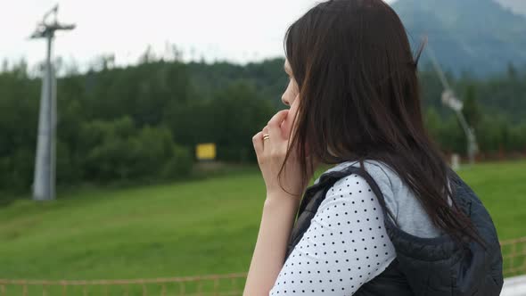 Woman in a Warm Vest Talking Smartphone Standing on the Background of Mountains and Moving Ski Lifts alt