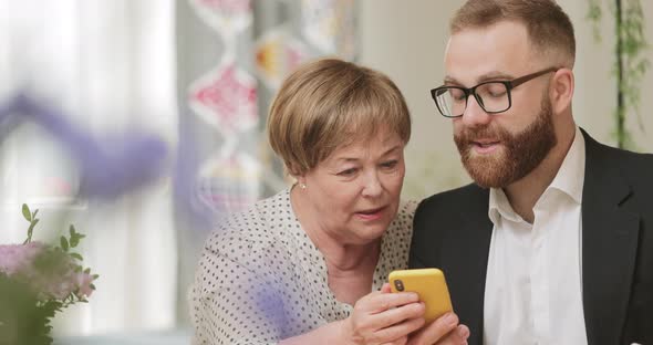 Cheerful Grandmother Andher Successful Son Sitting and Using Smartphone. Elderly Lady and Young Man alt