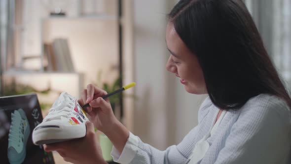 Female Footwear Designer Looking At The Laptop And Putting Coloured Unique Patterns On Sneakers alt