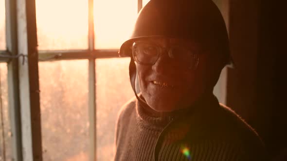 Elderly Man with Glasses Picks Up Military Helmet in Dust and Putting Helmet on Head in House Window alt