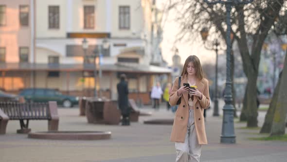 young attractive Woman in a warm coat walking on a City Street Using her Mobile Phone. alt