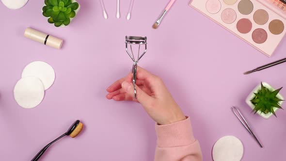 Female hand opens and closes metal eyelash curler on pink background with eyeshadow palette