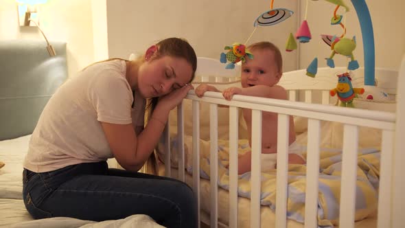 Little Baby Boy Shouting and Crying Next to Tired Mother Sleeping Next to His Crib alt