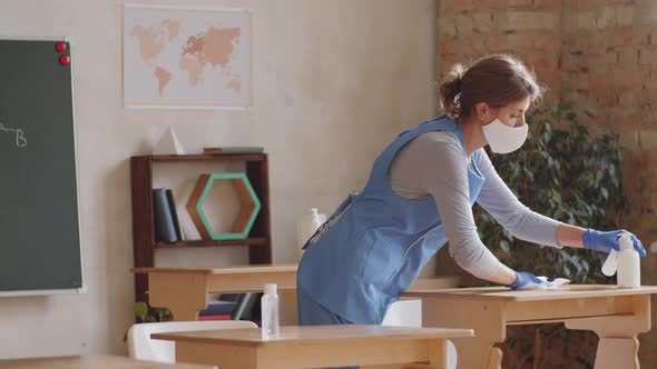 Female Cleaner in Mask and Gloves Disinfecting School Desks alt