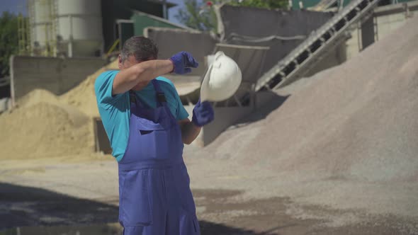 Portrait of Exhausted Factory Worker Taking Off Safety Helmet, Wiping Forehead with Hand, and alt