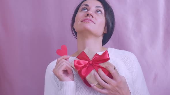 Smiling Funny Young Brunette in a White Sweater with a Gift and a Red Heart is Dancing on a Pink