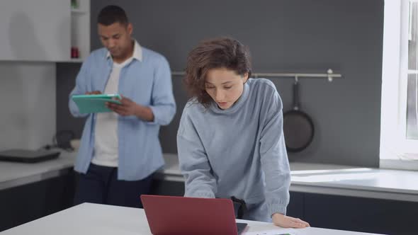 Focused Serious Caucasian Woman Typing on Laptop Keyboard Looking Back at African American Man alt