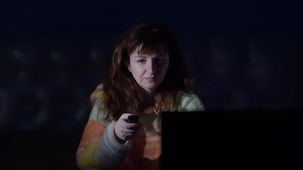 Brunette Woman Sitting on the Bed at Night and Watches TV Switches Channels with Different Emotions