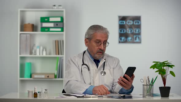 Aged Male Doctor Fills in Medical History and Advises Patient on Video Call Using Mobile Phone About alt