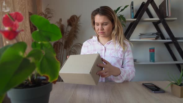 A Young Caucasian Lady is Sitting at the Table at Home and Opens a Box alt