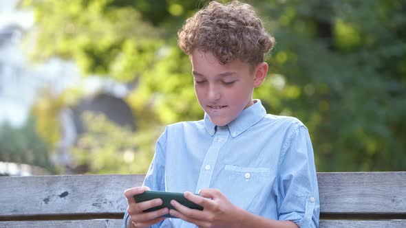 Young Boy Playing Game on His Smartphone Outdoors in Summer Park alt