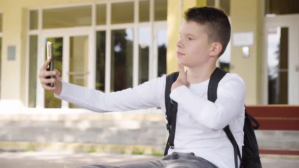 A Caucasian Teenage Boy Takes Selfies with a Smartphone  a School in the Background