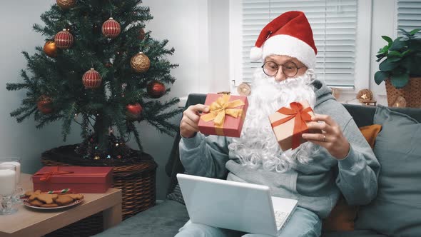 A Man in a Santa Claus Costume with a Beard Shows Gifts Via Video Link on a Laptop for Christmas to