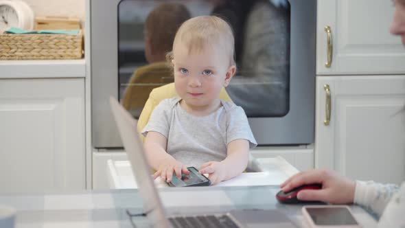 Mother Works on Laptop While Her Toddler Playing with Smartphone One Year Old Baby Boy Sitting in alt