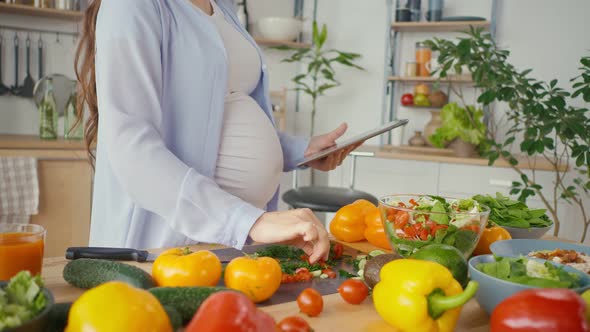 A Pregnant Woman Using a Tablet Prepares Healthy Food in a Bright Kitchen alt