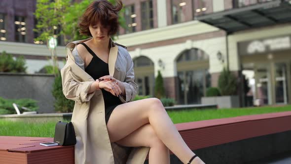 Woman Looks at Her Watch and Sits on a Bench in a Trench Coat