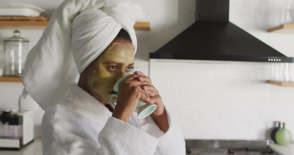 Happy african american woman with beauty mask on face, drinking coffee in kitchen alt