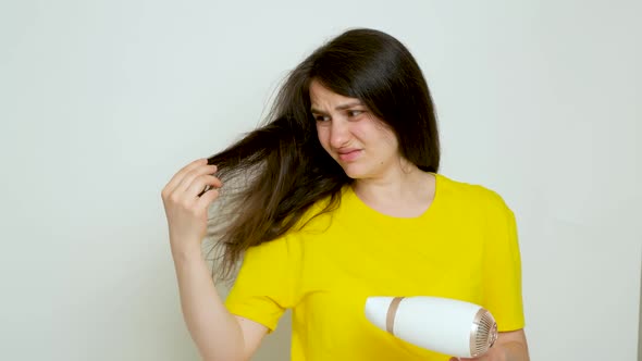 A Brunette Woman Dries Her Hair with a Hair Dryer Looks at Her Hair with a Dissatisfied Look alt