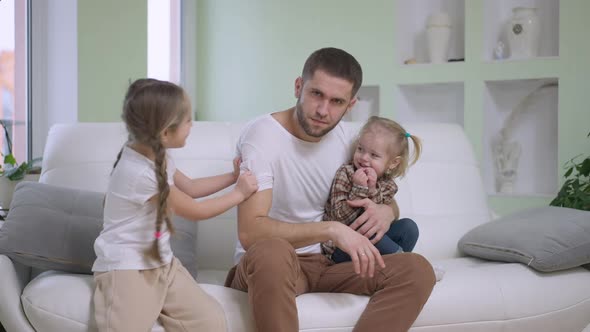 Exhausted Young Single Father Sitting on Couch Looking at Camera As Joyful Girls Tickling Man in alt