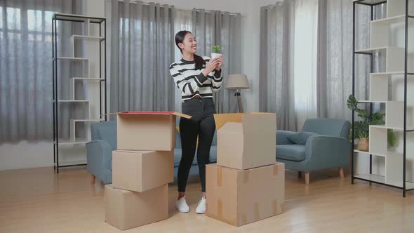 Young Asian Woman Moving Into A New House Unpacking The Cardboard Box And Taking Stuff Out Of It alt