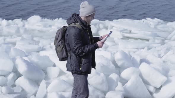 Young Blond Bearded Handsome Man in Warm Jacket and Hat Standing on the Glacier alt