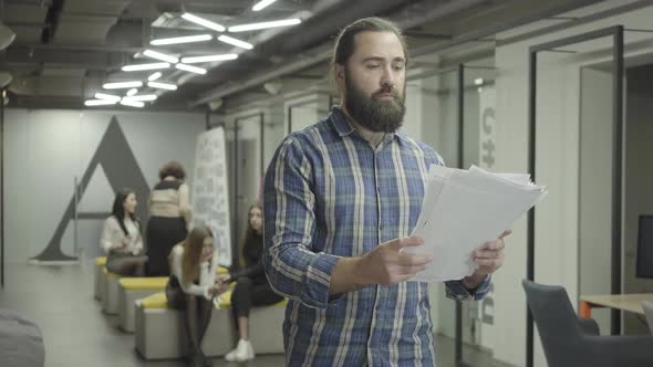 Concentrated Bearded Man Studying Papers Documents Standing in the Office. Young Colleague Riding alt