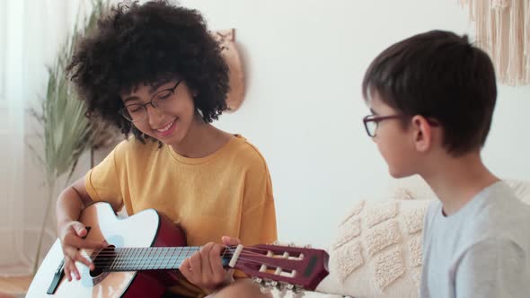 Afro American Woman Teaching Boy to Play Guitar While Sitting on Bed at Home alt