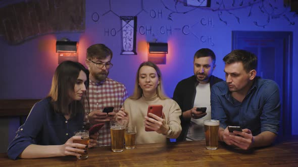 Friends in Pub at Bar Counter with Beer Look at Their Phones