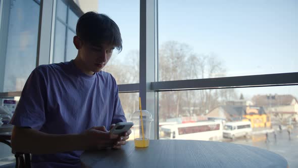 Young Guy is Sitting in a Cafe By the Big Window Drinking Juice and Having Fun on the Internet