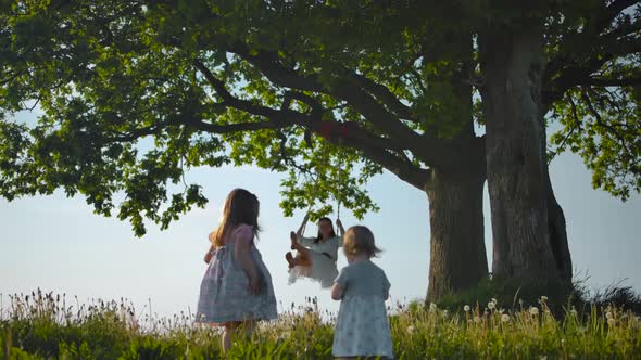 Sisters Walking Near Clean Green Field To Their Mother, Swinging on Swing Tied To an Old Oak-tree. alt
