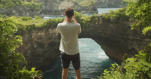 Back Shot of a Male Tourist Taking Pictures with His Phone Man Photographing Arch and Ocean Cliffs alt