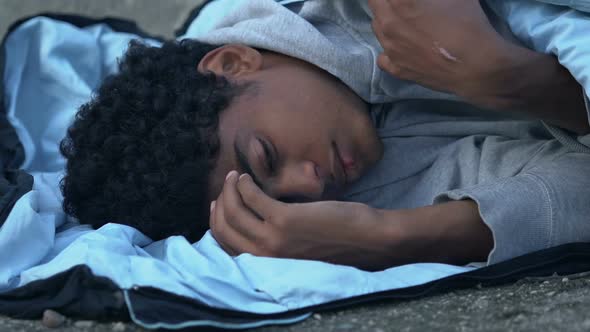 Freezing African-American refugee lying on street in sleeping bag alt