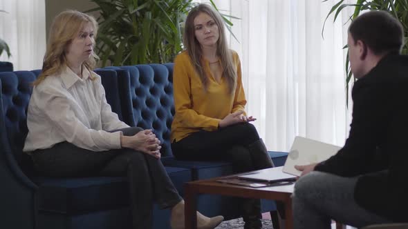 Portrait of Serious Caucasian Senior and Young Women Sitting on Couch and Listening To CEO alt