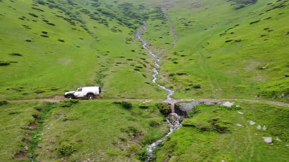A White Suv Rides Through A Mountainous Area Along A Dirt Road Past A River And A Waterfall.  Aerial