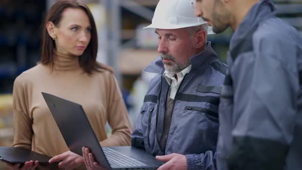 Professional Senior Man in Hard Hat Holding Laptop Discussing Shipping Plan with Young Employees alt