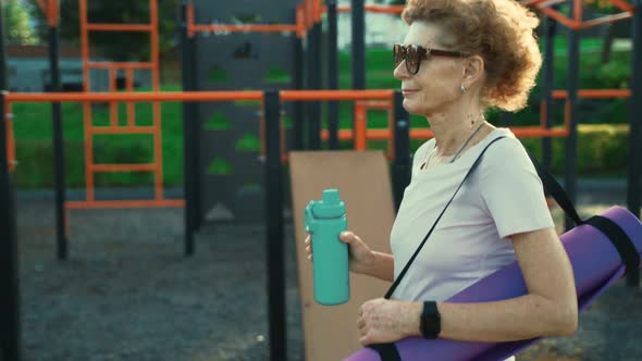 Active Senior Woman with Fitness Mat and Water Bottle in Hands Walking on Workout Outdoor Gym alt