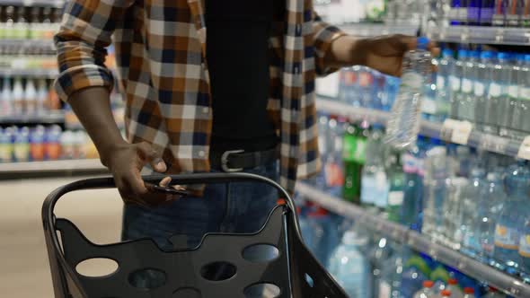 Unrecognizable Man with Smartphone Pushes Shopping Cart Taking Bottle alt