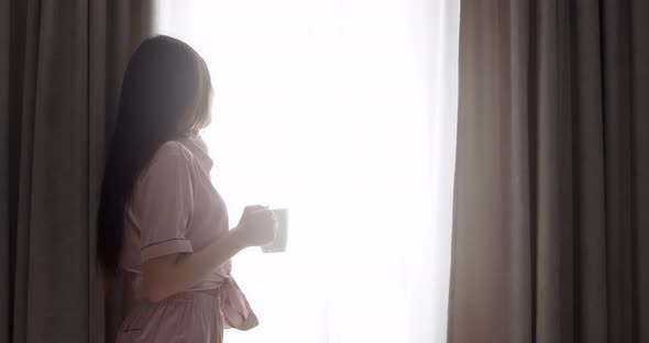 Woman is Talking on a Cell Phone Near the Window and Drinking Tea From Mug