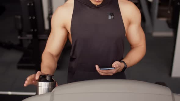 Man of Athletic Build with Sports Bottle and Smartphone in Hand Approaches Treadmill and Starts alt