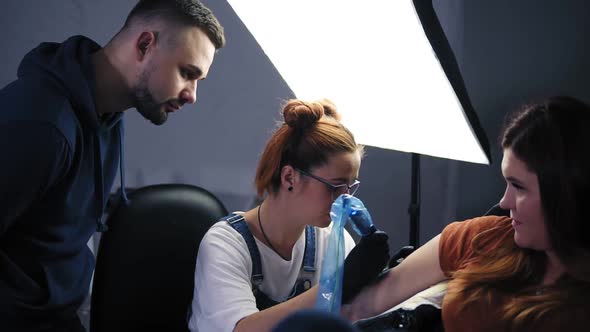 The Girl  the Master of Tattoos Providing a Lesson for Man Standing Above and Watching the Process alt