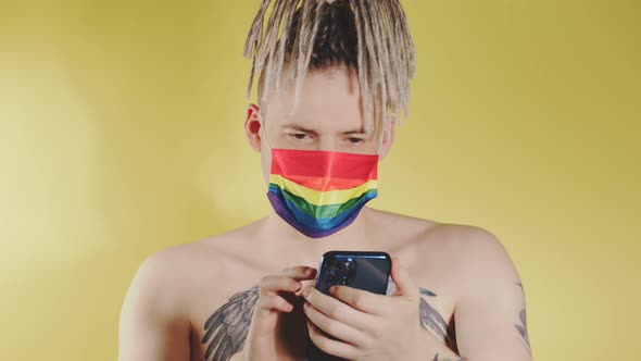 Naked concentrated man in LGBT mask browsing mobile phone. Focused gay guy with blonde dreadlocks alt