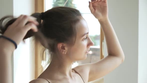 Sporty Brunette Pretty Female Stands at Window Fixes Hair Collects Hair in Bun